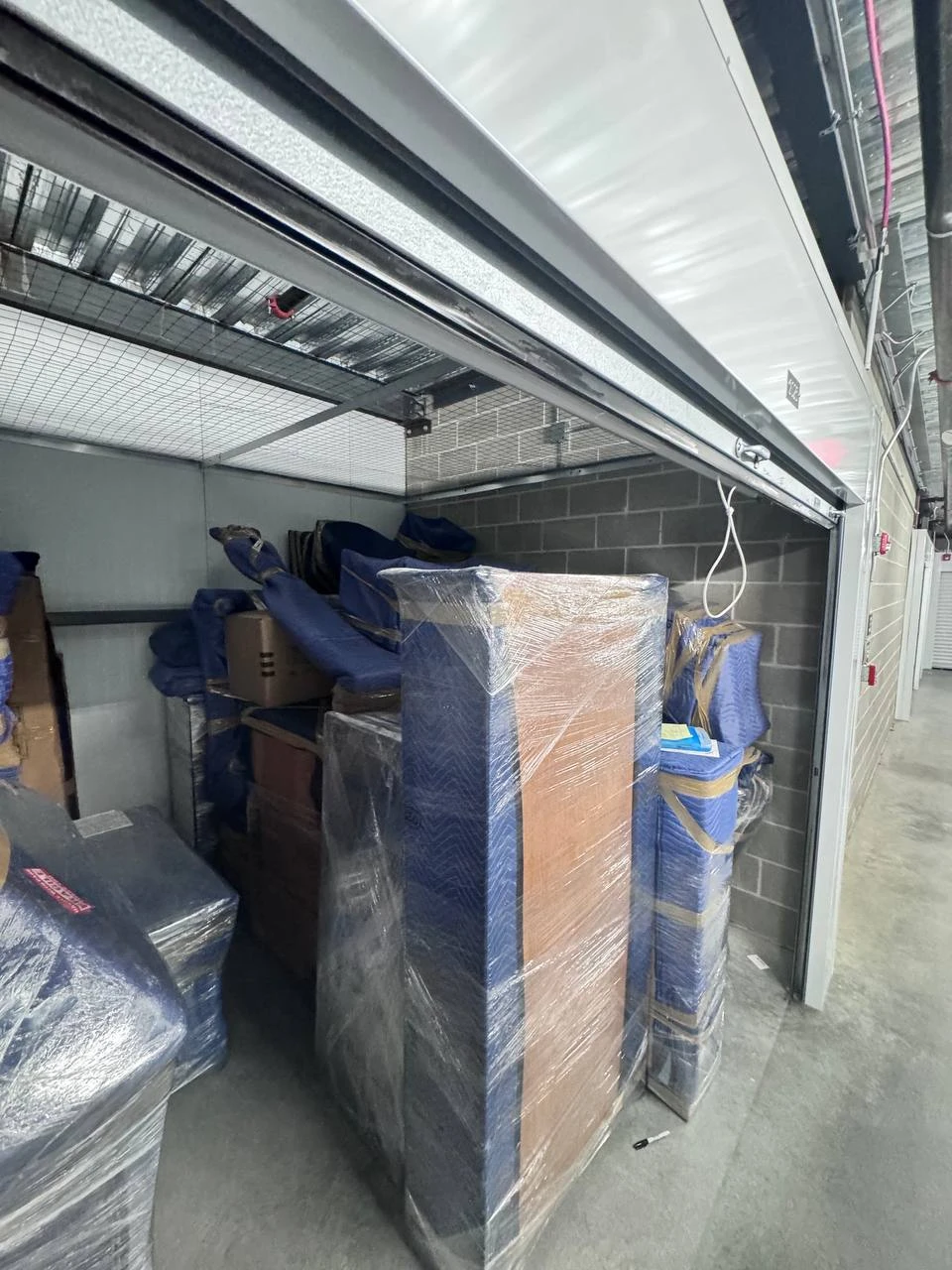 Inside a Raimond's Movers storage unit with wrapped furniture, mattresses, and boxes neatly organized behind the roll-up door